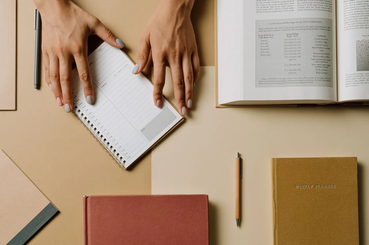 Hands opening a spiral daily planner on a beige desk beside notebooks and an open book